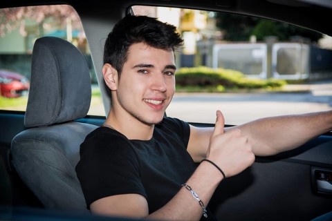 Happy young man or teenager doing thumb up sign while driving car and smiling at camera