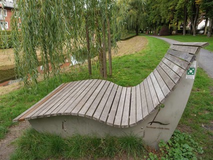 A curved wooden bench stands under a willow tree along a park path, suitable for relaxation,