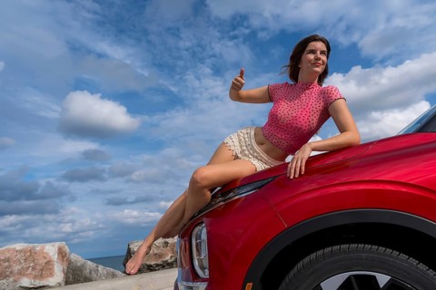 A gorgeous redhead model sits confidently on the hood of her car, soaking in the summer breeze as