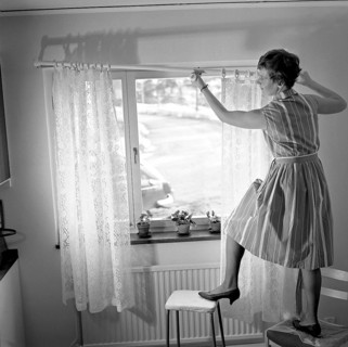 1960s home decorating. A woman at home putting up the curtains in the room window. All by herself
