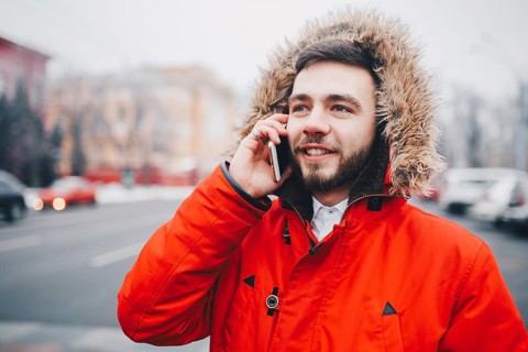 Young guy with beard and red jacket in hood a student uses mobile phone, holds in his hand near the