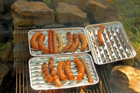 Grilled sausages lie on an aluminium grill tray over a campfire