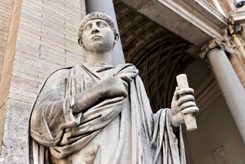 Historical marble statue of a person holding a scroll in front of a classical column structure,