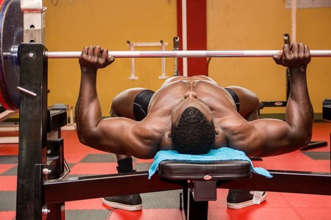 Hunky muscular black bodybuilder working out in gym, exercising pecs on bench with barbell