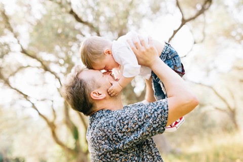 The father holds his son above him, they touching their noses, the son holds the father by the