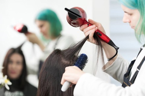 Female hairstylist dries brunette hair with red hair dryer and blue hairbrush in professional