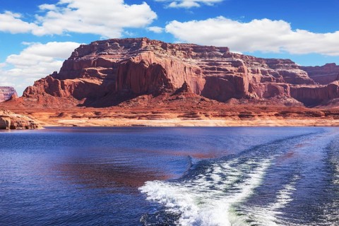 Foamy trace of motorboat crosses the emerald waters. Red sandstone hills surround the lake. Lake