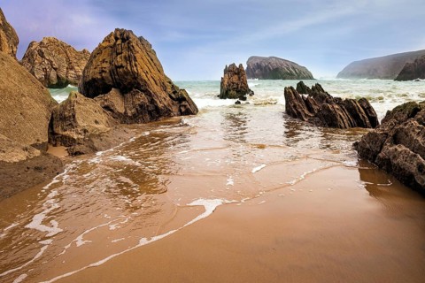 The smooth wet sand of the beach reflects the rocky boulders. The Playa de la Arnia beach.