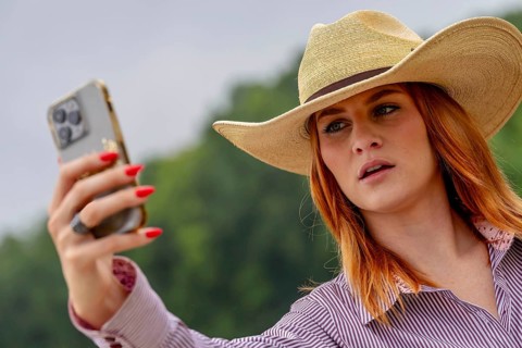 A beautiful redhead model uses her cellphone while posing in a country setting wearing cowgirl
