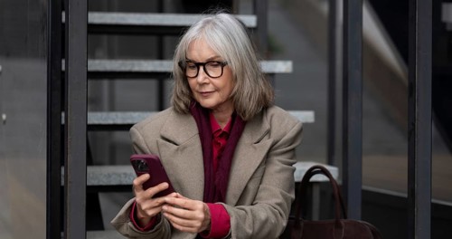 A middle-aged businesswoman dressed in a stylish coat and glasses is engaged with her smartphone