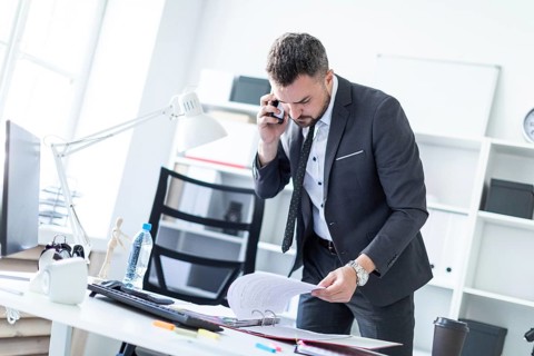 A man is standing near a table in the office, talking on the phone and flipping through documents