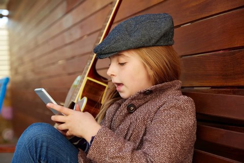 Blond kid girl playing smartphone winter beret and guitar on wooden background