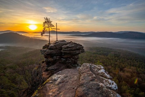 Sunrise on a rock with a summit cross and a view over a forest. Morning atmosphere with fog in the