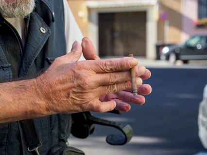 Older caucasian man sits on medical walker outdoors, smoking a cigar in public despite visible