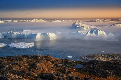 Large glaciers in the bay by the rocky shore in the light of the setting sun with fog over the
