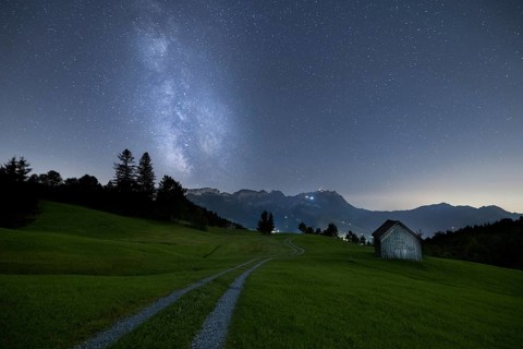 A clear night with a view of the Milky Way over a meadow and a remote hut with a mountain view,