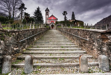 long stairs leading up to an old rose colored church under an expressive overcast sky in the Maggia