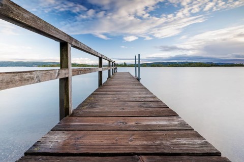 Long wooden footbridge leads into a quiet lake under a partly cloudy sky, Naturfreundehaus,