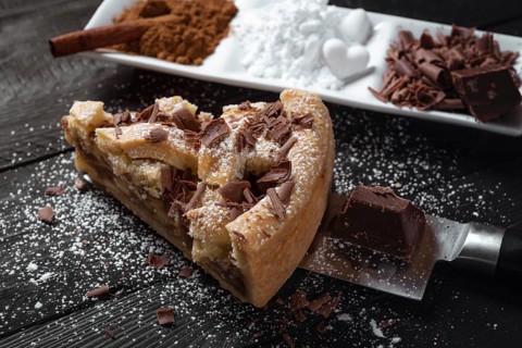 Slice of apple pie with powdered sugar and chocolate, on a knife, on a black rustic table and a