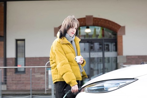 Woman in yellow jacket holding a cup, standing by charging electric car in urban environment,