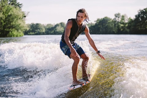 Young athletic man with long hair wakesurfing on waves of river in sunny summer weather. Ttheme