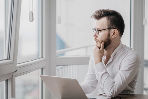Pensive man in glasses and a white shirt looking out of a window beside his laptop