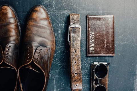 Flat lay of shoes, belt, passport, and sunglasses arranged on a dark scratched surface