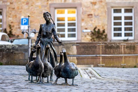 Statue of a girl with geese in front of a historic building, a parked car in the background, The