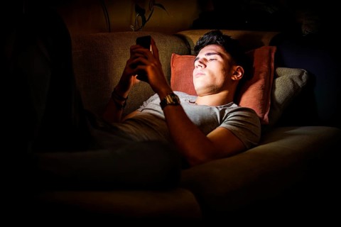 Attractive Young Man Lying on Sofa at Night and Illuminated by Light from Screen of Cell Phone at