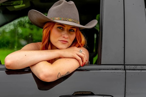 A beautiful redhead model sits in her truck while wearing a cowboy hat on her farm