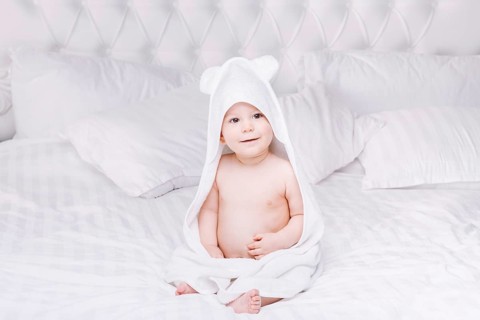 Adorable newborn baby boy with brown eyes sitting wrapped in a white towel with ears in light bed.