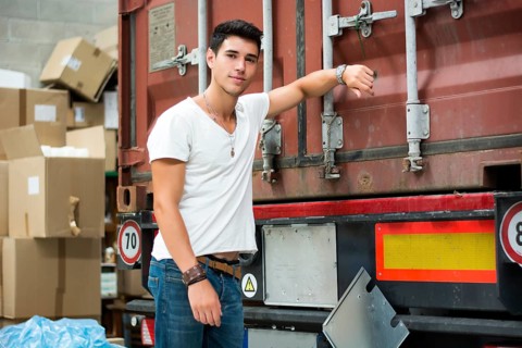 Waist Up Portrait of Young Smiling Man Next to Freight Truck Leaning against it, Looking at Camera