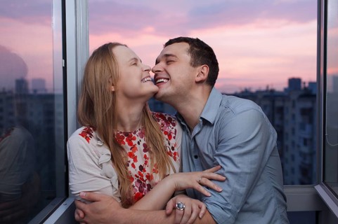 Playful young couple embracing and laughing on the balcony at a high floor during sunset