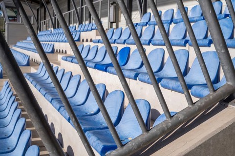 Rows of empty blue plastic stadium seats behind a metal barrier await the return of fans