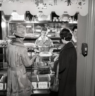 1960s coffee shop. Two women are standing in front of the counter that is full with freshly baked