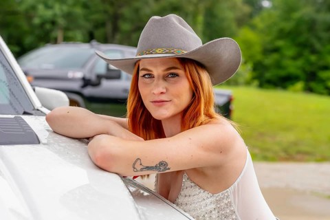 A beautiful redhead model poses in a country setting wearing cowgirl attire against her truck