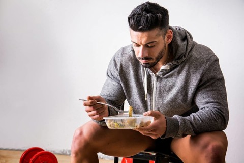 Hungry muscular young man gulping down food looking at plastic container without pausing as he