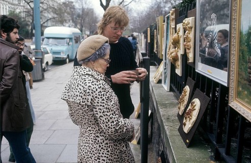 London 1982. A view from a London street with an elderly lady looking at art hanged on the fence to