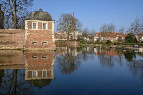 A pavilion at the moat with a clear reflection, surrounded by trees in the park in front of a blue