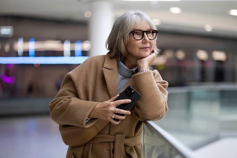 A woman with stylish glasses leans against a railing, thoughtfully holding her smartphone. She
