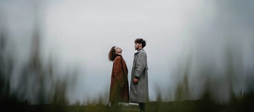 A couple standing in a field, each looking upwards, dressed in long coats with a sense of