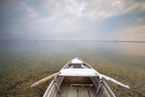 A snow-covered boat on a calm lake with pebbles on the shore under a cloudy sky, Strandbad Hörnle,