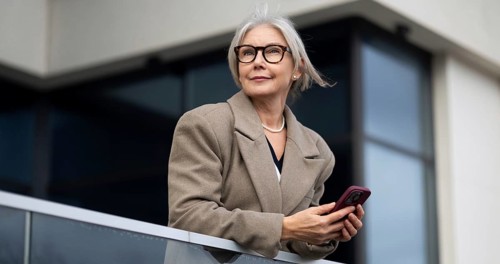 The stylish elderly woman stands elegantly on a balcony, holding her phone and gazing thoughtfully