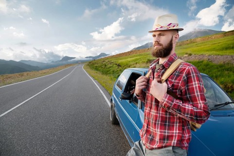 Attractive bearded man in a plaid red shirt and hat with a backpack. Stands next to a retro car on