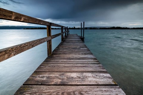 A long wooden footbridge leads into the water of a lake under a dark, cloudy sky, Naturfreundehaus,