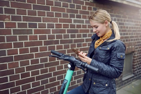Trendy young blond woman in leather jacket standing at her e-scooter booking a ride, ecological