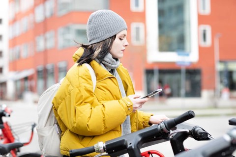 Woman in yellow jacket scrolling on her smartphone next to parked rental bike in the city,