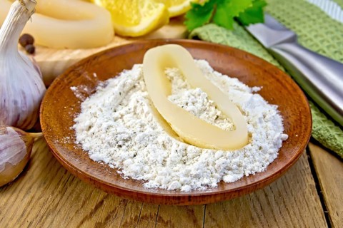 Raw squid rings in flour on a plate, garlic, knife, lemon, napkin on the background of wooden