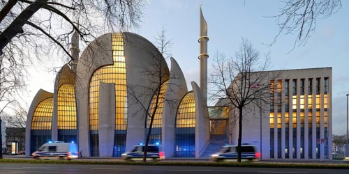 Several police vehicles with flashing lights drive past the Kölne Mosque in Ehrenfeld