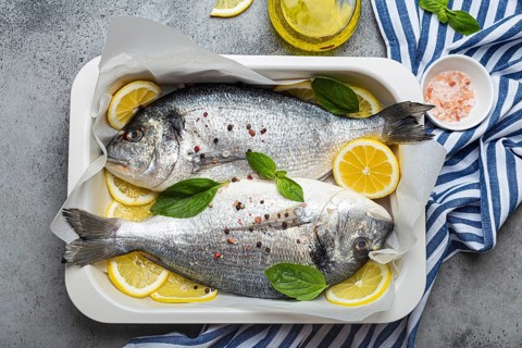 Raw fish dorado in casserole dish with ingredients lemon, fresh basil, bottle of olive oil on